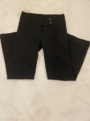 Svarta kostymbyxor med hög midja - Stilrena svarta kostymbyxor med hög midja och två knappar framtill. Byxorna har raka ben och är sydda i ett mjukt, följsamt tyg som ger en dressad look. Perfekta för en trendig och clean outfit.