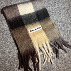 Rutig halsduk från Acne Studios - Mjuk och fluffig halsduk från Acne Studios i rutigt mönster med nyanser av beige, brunt, grått och vitt. Halsduken har långa fransar i ändarna och en tydlig Acne Studios-logga. Perfekt accessoar för att lyfta din outfit. Pris kan diskuteras 