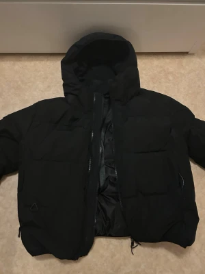 Svart pufferjacka från ZARA - Säljer en svart pufferjacka från ZARA med huva och dragkedja framtill. Jackan har två sidofickor med dragkedja och justerbar nederkant. Insidan har praktiska remmar för att bära jackan över axeln. Perfekt för kalla vinterdagar.