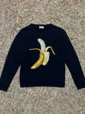 Svart sweatshirt Acne Studios Banana - Cool svart sweatshirt från Acne Studios med ett stort broderat bananmotiv på bröstet. Tröjan har rund hals och långa ärmar, perfekt för en avslappnad och trendig look. Lite urtvättad bara och där med kan priset också diskuteras. 