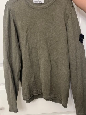 Olivgrön tröja från Stone Island - Snygg olivgrön långärmad tröja från Stone Island med klassisk patch på vänster ärm. Tröjan har rund halsringning och är tillverkad i ett mjukt, tunt stickat material som ger en avslappnad look. Perfekt för dig som gillar streetwear och stilrena plagg.