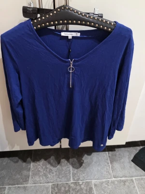 Blå långärmad topp från Flash Woman - Mörkblå långärmad topp från Flash Woman med v-ringning och dekorativ dragkedja framtill. Tillverkad i mjukt material som känns skönt mot huden. Perfekt för dig som gillar enkel men snygg stil.