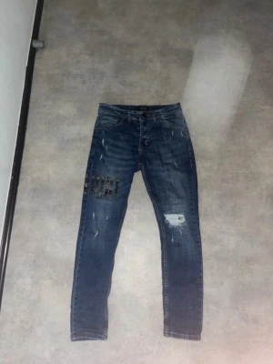 Mörkblå AMIRI jeans med patch - Snygga mörkblå jeans med skinny fit och slitningar på flera ställen. Ena benet har en cool patch med rutigt mönster. Klassisk femficksmodell och normal midja. Perfekta för en trendig streetstil.