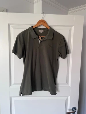 Olivgrön pikétröja från Burberry - Klassisk olivgrön pikétröja från Burberry med korta ärmar och krage. Tröjan har diskret logga broderad på bröstet och typisk Burberry-detalj vid knappslån. Perfekt till jeans eller shorts.