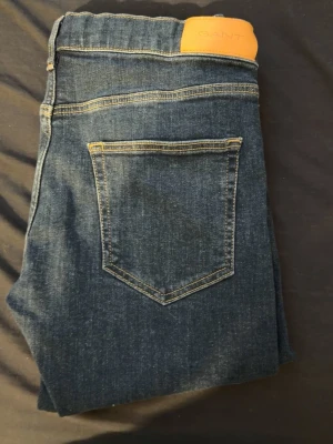 Mörkblå jeans från GANT - Snygga mörkblå jeans från GANT med klassisk femficksdesign och kontrastsömmar. Jeansen har en läderpatch med logga bak i midjan och är tillverkade i ett mjukt denimtyg. Perfekta för en stilren och avslappnad look.