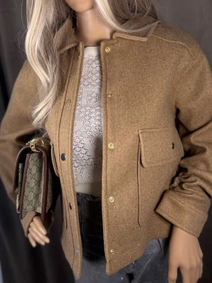 Beige overshirt med ficka M-L☑️☑️☑️ - Stilren beige overshirt i ullmix med klassisk krage och tryckknappar i guld. Jackan har en stor bröstficka med lock och raka ärmar. Perfekt lager-på-lager-plagg för en trendig look.
