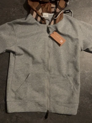 Grå hoodie från Burberry med dragkedja - Snygg grå hoodie från Burberry med hel dragkedja och klassiskt rutmönstrat foder i huvan. Hoodien har två fickor framtill och ribbade muddar. Perfekt för en stilren och avslappnad look.