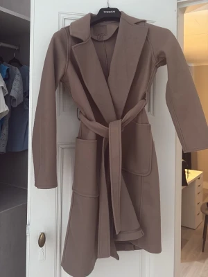 Brun kappa med knytskärp från new yorker - Stilren brun kappa från new yorker med knytskärp i midjan och stora fickor, lång ärm. Lite nopprig 