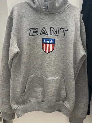 Grå hoodie från GANT med logga - Snygg grå hoodie från GANT med stor broderad logga och amerikanskinspirerat sköldmärke på bröstet. Är väldigt bekväm som en varje dag stil. Har XXL som size men passar även mig som M/L size. Har ljusare färg i verkligheten!