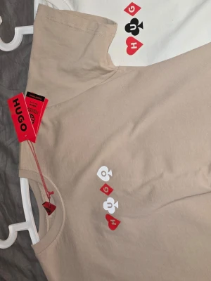 Beige HUGO t-shirt med design - Snygg beige t-shirt från HUGO med tryck av spelkort och texten 'Full House' på bröstet. Framsidan har även små symboler i rött, vitt och svart. Klassisk rund hals och korta ärmar. Perfekt för dig som gillar unika prints och streetstyle.