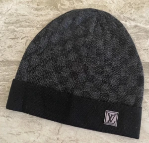 Svart  mössa med rutmönster - Snygg svart mössa från diskret rutmönster och vikt kant. Märkets klassiska logga sitter på en patch framtill. Perfekt accessoar för dig som vill ha en stilren och exklusiv look.