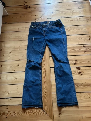 Blå True Religion  jeans med slitningar - Säljer ett par blå bootcut jeans med slitna detaljer på knäna och råa kanter nertill. Jeansen har klassisk femficksdesign och dragkedja med knapp. Perfekta för en avslappnad och trendig look.