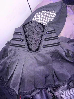 Svart kort korsettkjol från Killstar - Säljer en svart kort korsettkjol från Killstar med dekorativa broderier och knappar framtill. Kjolen har en plisserad kjol, spetsdetaljer och en edgy goth-vibe. Perfekt för dig som gillar alternativ stil. Saknas en knapp men finns en extra att sy på! 