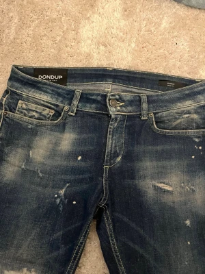 Dondup George jeans  - Säljer ett par blå jeans från Dondup med cool slitning och färgstänk för en trendig look. Jeansen har fem fickor, smal passform och klassisk knappgylf. Perfekta för dig som gillar en avslappnad men ändå stilsäker vibe.