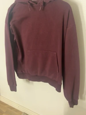 Vinröd hoodie med huva - En enkel och stilren vinröd hoodie med huva och dragsnören. Hoodien har en stor magficka framtill och ribbade muddar vid ärmslut och nederkant. Perfekt för en avslappnad och chill look. Materialet känns mjukt och bekvämt, troligen bomull eller bomullsblandning.