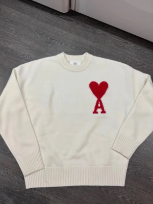 Vit stickad tröja från Ami Paris - Vit stickad tröja från Ami Paris med röd hjärtlogga och bokstaven A på bröstet. Tröjan har rund halsringning, ribbade muddar och är tillverkad i mjukt material. Perfekt för en stilren och trendig look.