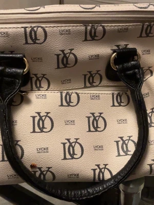 Beige handväska från Lycke Oslo - Snygg beige handväska från Lycke Oslo med svart logotypmönster över hela väskan. Väskan har svarta handtag i konstläder och guldfärgade metalldetaljer. Klassisk form med dragkedja upptill och stilren design som sticker ut.