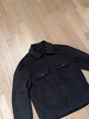 Mörkblå overshirt från Zara - Stilren mörkblå overshirt från J.Lindeberg med klassisk krage och fyra framfickor med lock och knapp. Overshirten har raka ärmar och knäppning framtill. Materialet känns kraftigt och passar perfekt för lager-på-lager.