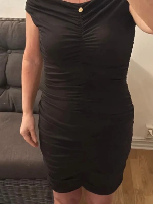 Svart offshoulder kort klänning - Snygg svart kort klänning med offshoulder-design och rynkade detaljer framtill. Klänningen har en tight passform och är perfekt för dig som vill ha en stilren och elegant look. Tillverkad i ett stretchigt material som sitter bekvämt. Passar S/M, reglerbar längd.