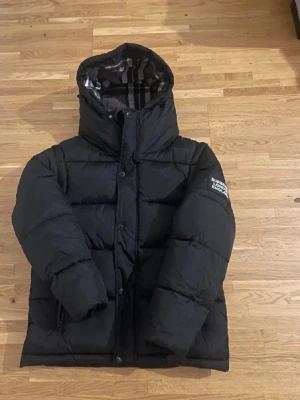  Burberry jacka  - Säljer en svart pufferjacka från Burberry med klassiskt rutigt foder i huvan. Jackan har hög krage, dragkedja och knappar framtill samt två fickor med dragkedja. Perfekt för kalla vinterdagar och riktigt snygg streetstil.