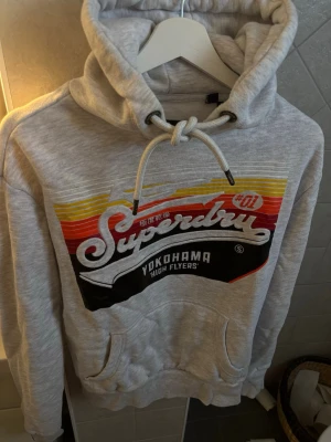Grå Superdry hoodie med tryck - Snygg gråmelerad hoodie från Superdry med färgglatt retrotryck i rött, gult och svart framtill. Stor logga och texten 'Yokohama High Flyers' på bröstet. Klassisk huva med dragsko och känguruficka. Perfekt för en avslappnad streetwear-look.