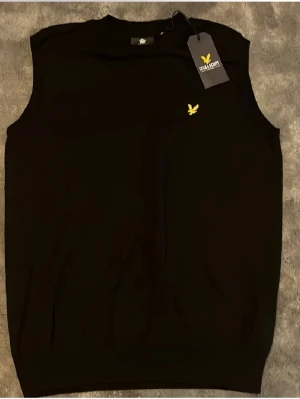 Svart väst från Lyle & Scott - Svart väst från Lyle & Scott med klassisk rund hals och gul logga broderad på bröstet. Västen är enkel och stilren, perfekt att bära över en skjorta eller t-shirt. Materialet känns mjukt och bekvämt.🤩
