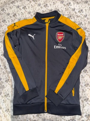 Arsenal träningsjacka Puma grå/gul - Snygg Arsenal träningsjacka från Puma i mörkgrått med gula detaljer längs ärmar och rygg. Jackan har dragkedja, ribbade muddar och Arsenal-logga samt Fly Emirates-tryck på bröstet. OBS. STORLEKEN ÄR 12Y