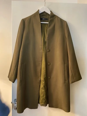 Olivgrön kappa från Gina Tricot - Stilren olivgrön kappa från Gina Tricot med rak passform och tre knappar framtill. Kappan har diskreta fickor på sidorna och är fodrad i matchande färg. Perfekt för dig som gillar minimalistisk och trendig look.