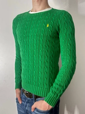 Grön kabelstickad tröja från Ralph Lauren - Snygg grön kabelstickad tröja från Ralph Lauren med gul broderad logga på bröstet. Tröjan har rund halsringning, långa ärmar och ribbade muddar. Perfekt för dig som gillar klassisk och stilren design.