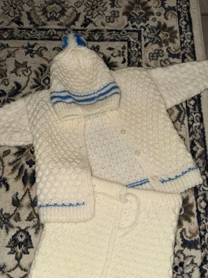 Vit stickad babyset med blå detaljer - Säljer ett handstickat set för baby i mjuk vit ull med blå ränder. Setet består av en kofta med knappar, byxor med dragsko och en mössa med blå ränder och tofs. Perfekt för kyliga dagar och supergulligt på små barn.