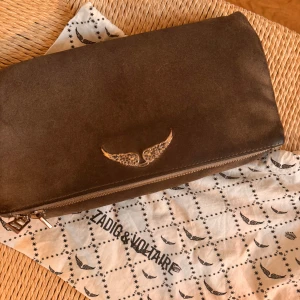 Grå mocka kuvertväska Zadig & Voltaire - Säljer en brun kuvertväska i mocka från Zadig & Voltaire med snyggt vingemblem i metall framtill. Väskan har dragkedja och stilren design, perfekt för att lyfta din outfit. Kommer med originalpåse med logga.pris kan diskuteras 💕