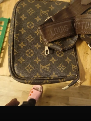 Louis Vuitton Monogram axelväska - Snygg axelväska från Louis Vuitton i klassiskt brunt monogrammönster med beige detaljer. Väskan har justerbar axelrem i brunt tyg med Louis Vuitton-logga och en rund liten myntplånbok som hänger på remmen. Tillverkad i canvas med läderdetaljer.