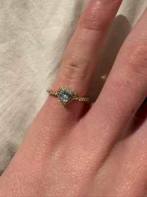 Guldfärgad ring med hjärtformad sten - Snygg guldfärgad ring med en stor hjärtformad klar sten i mitten, omgiven av små gnistrande stenar runt hela hjärtat och längs ringen. Ringen har en elegant och glittrig design som verkligen drar blickarna till sig. Perfekt för dig som gillar romantiska detaljer och bling.