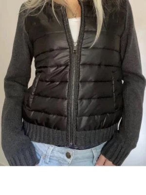 Snygg cardigan  - Snygg svart pufferjacka med quiltad front och rygg i glansigt material samt stickade, mörkgrå ärmar och muddar. Jackan har dragkedja framtill och två fickor. Perfekt för kalla dagar när du vill vara både varm och stilren.