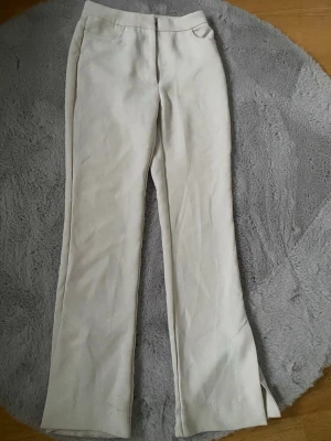Beiga kostymbyxor med slits - Stilrena beiga kostymbyxor med raka ben och diskret slits nertill. Byxorna har hög midja. Perfekta att matcha med en skjorta eller stickad tröja. Materialet känns mjukt och följsamt.