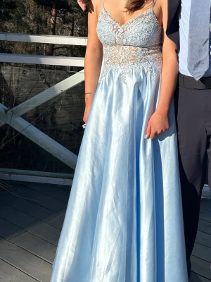 Ljusblå långklänning med spetsdetaljer - Elegant ljusblå balklänning med tunna axelband och v-ringad urringning. Överdelen har vackra spetsdetaljer och transparent parti i midjan. Kjolen är utsvängd och tillverkad i glansigt material som ger en lyxig känsla. Perfekt för bal eller speciella tillfällen. Använd en gång, och uppsydd så passar mig perfekt som är 164 och som hade klackar. Original pris 2500