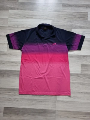 Svart och rosa pikétröja med gradient - Snygg pikétröja med svart till rosa gradient och korta ärmar. Tröjan har krage, knappar framtill och ett litet broderat djurmotiv på bröstet. Perfekt för dig som gillar färg och sportig stil.