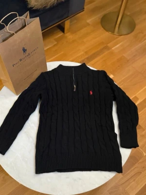 Svart kabelstickad tröja Polo Ralph Lauren - Kabelstickad svart tröja från Polo Ralph Lauren med halv dragkedja och ribbad krage. Tröjan har det klassiska röda Polo-logotypen broderad på bröstet och är tillverkad i mjukt material. Perfekt för lager på lager och en stilren look.