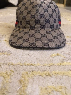 Gucci beige keps med GG-mönster - Säljer en beige keps från Gucci med det klassiska GG-monogrammet över hela kepsen. På sidan finns en ikonisk röd och marinblå rand. Kepsen har böjd skärm och justerbar rem bak. Materialet är canvas med detaljer i tyg.