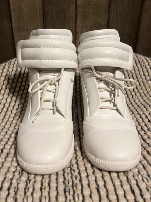Maison Margiela futures - Säljer ett par vita höga sneakers från Maison Margiela. Skorna har en chunky siluett, bred vadderad plös och krage samt vita snören. Tillverkade i slätt läder för en clean och futuristisk look. Perfekta för dig som gillar statement pieces och exklusiv design. Skriv för frågor eller mer bilder. Pris går att diskutera
