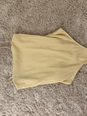 Ljusgul ribbad linnetopp - Säljer en ljusgul ribbad linnetopp med tunna axelband. Toppen har en enkel och stilren design med smala ränder och är perfekt för varma dagar. Materialet känns mjukt och följsamt mot huden.