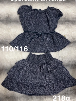 Mörkblått set med topp och kjol - Tvådelat set i mörkblått med små vita prickar. Toppen har korta puffärmar, resår vid halsen och ett matchande midjeband. Kjolen är vid och har resår i midjan. Perfekt för en trendig och bekväm look.🌸Sparsamt använda. Storlek 110/116 