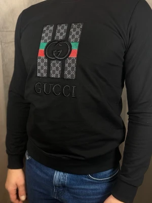 Svart långärmad topp f - Svart långärmad topp  med broderad logga och klassiska  ränder i grönt och rött framtill. Toppens material känns mjukt och stretchigt, perfekt för en trendig look. Snygg detalj och ribbade muddar vid ärmslut och midja