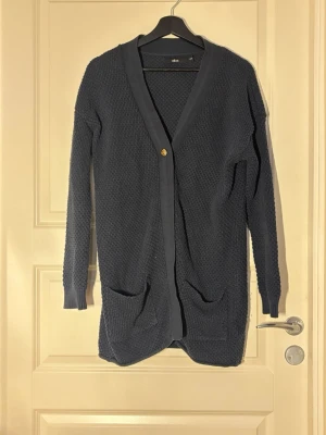 Mörkblå lång kofta med fickor - Mysig mörkblå stickad kofta från ellos med lång passform, djupa fickor framtill och en enkel knappstängning. Koftan har ribbade muddar och en grovstickad struktur som ger en avslappnad look. Perfekt att slänga på sig över en t-shirt eller topp.