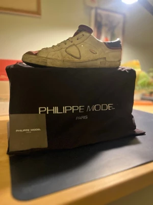 vinröd Philippe Model sneakers - Snygga vit sneakers från Philippe Model med bruna och vinröda detaljer. Skorna har en klassisk låg siluett, rund tå och är tillverkade i mocka och läder. Perfekta för dig som gillar en stilren men ändå unik look. Nypris ca 5000