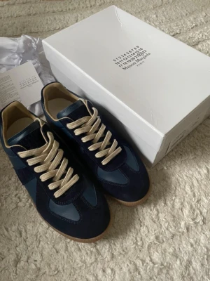 Maison Margiela  - Snygga sneakers från Maison Margiela i mörkblå mocka och läder med beige snörning och gummisula. Klassisk low-top modell med stilren design och diskreta detaljer. Perfekta för dig som gillar exklusiva och trendiga skor.