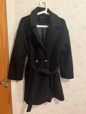 Svart kappa med bälte från Vero Moda - Stilren svart kappa från Vero Moda med klassisk krage, dubbelknäppning och avtagbart bälte i midjan. Kappan har slät yta och är helfodrad, perfekt för kyligare dagar. Enkel men trendig design som passar till många outfits.