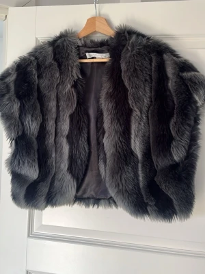 Mörkgrå pälsväst i fuskpäls - Snygg mörkgrå pälsväst i fuskpäls med kort ärm och öppen front. Västen har ett lyxigt, fluffigt utseende och är fodrad på insidan för extra komfort. Perfekt för att lyfta din outfit med en trendig touch.