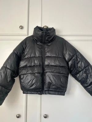 Svart cropped pufferjacka - Snygg svart pufferjacka i cropped modell med hög krage och dragkedja framtill. Jackan har breda kanaler och elastisk nederkant för extra komfort. Perfekt för kalla dagar när du vill hålla stilen. Materialet är glansigt och ger en modern look.