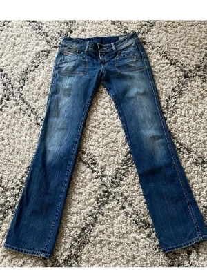 Blå bootcut jeans med broderi - Säljer ett par blå bootcut jeans med snygga slitningar och orange broderi på bakfickorna. Jeansen har klassisk femficksmodell och detaljerad söm. Perfekta för dig som gillar en avslappnad men ändå trendig look.